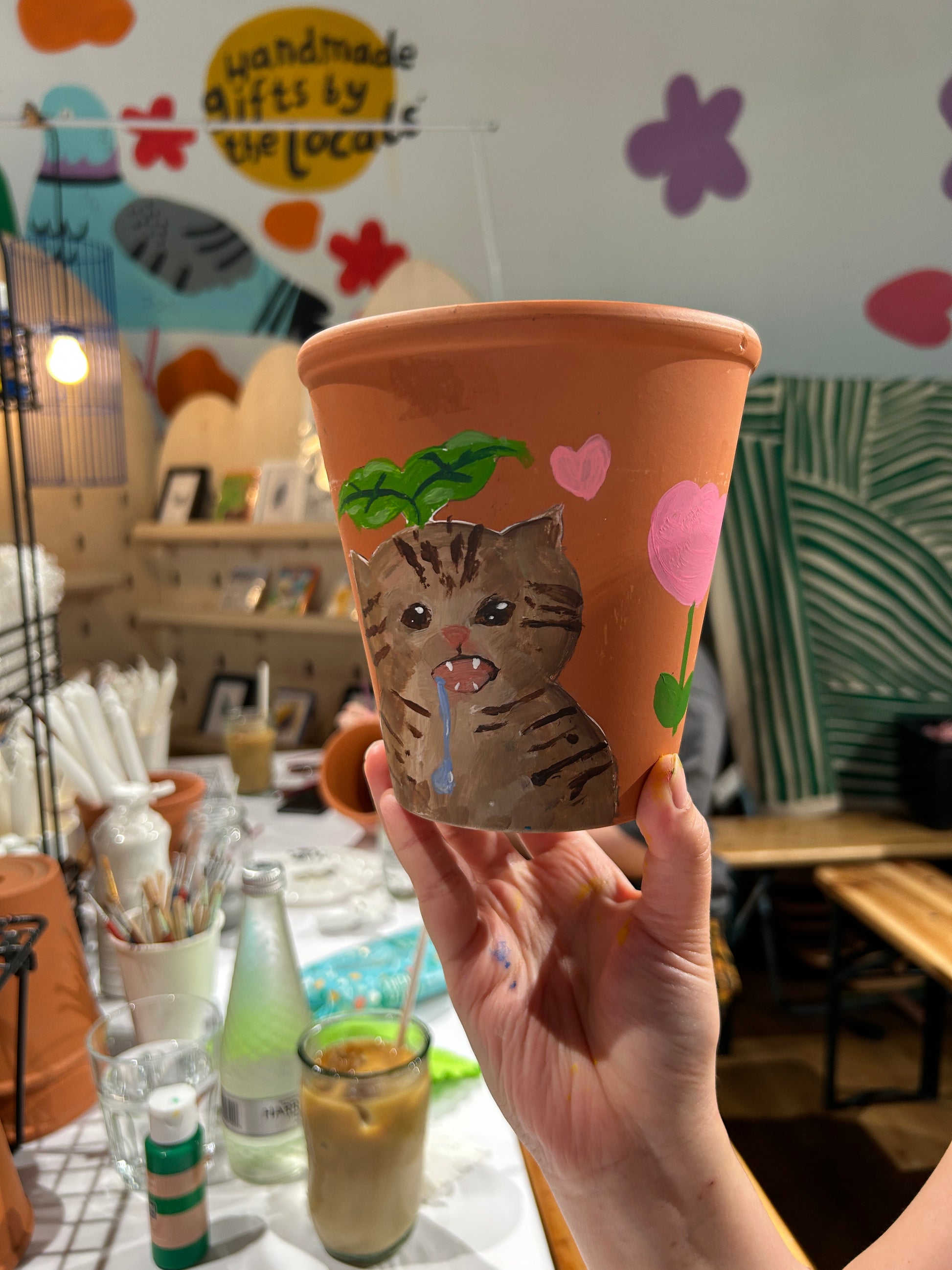 A hand holding a terracotta-colored paper cup decorated with a hand-painted brown tabby cat, pink hearts, and green leaves, set against a busy cafe background.